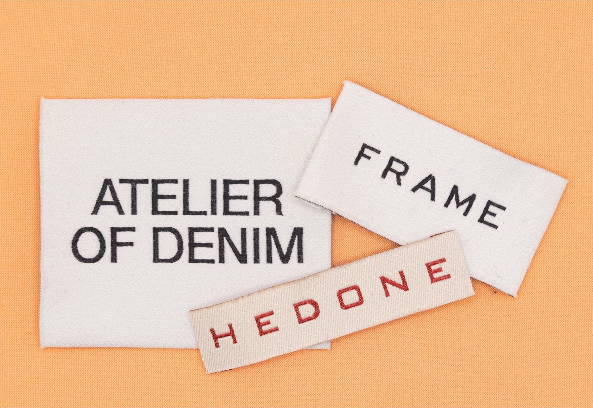 A collection of white woven cotton labels for brands like 'ATELIER OF DENIM', 'FRAME', and 'HEDONE' on an orange background.